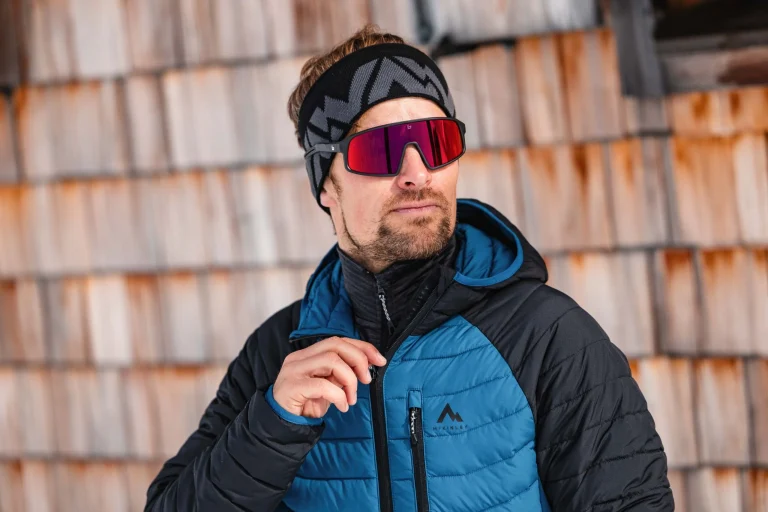 Mann mit roter Skibrille und Winterjacke vor Holzwand