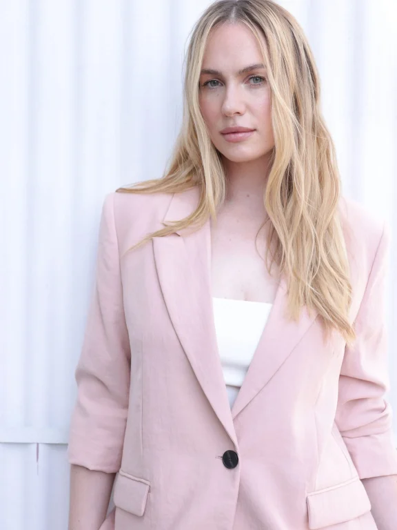 Blonde Frau im rosafarbenen Blazer vor weißer Wand