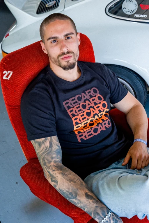 Tätowierter Mann im Rennsportshirt vor weißem Sportwagen
